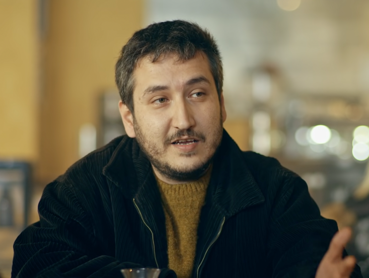 A film still of a bearded man in a corduroy jacket speaking across a café table, used here as a reaction shot